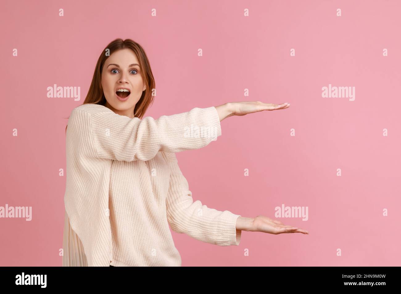 Porträt der aufgeregt erstaunt schöne blonde Frau präsentiert Bereich zwischen den Händen für Werbung, zeigt riesige Größe, trägt weißen Pullover. Innenaufnahme des Studios isoliert auf rosa Hintergrund. Stockfoto