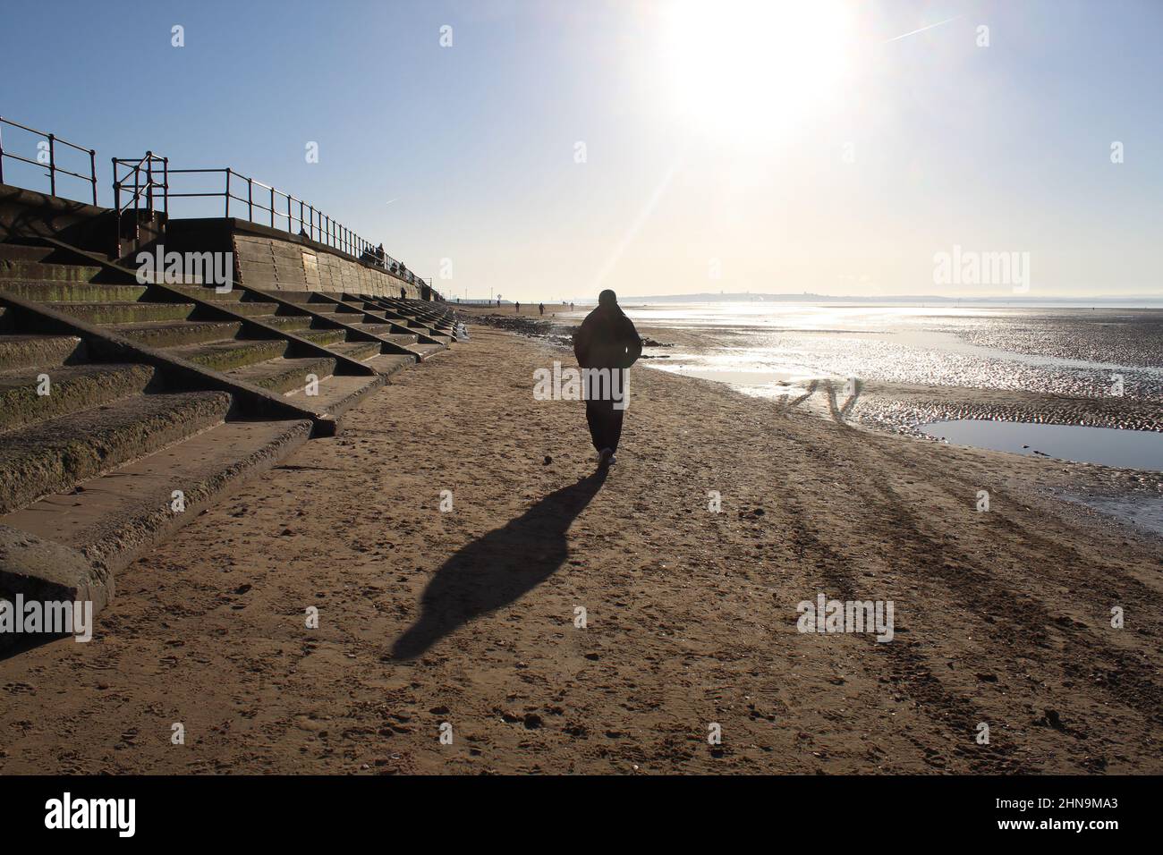 Ein Mann, der an einem kalten Morgen in Richtung Sonne am Strand geht. Er ist allein und denkt über seine Gedanken nach, gut für seine körperliche und geistige Gesundheit Stockfoto