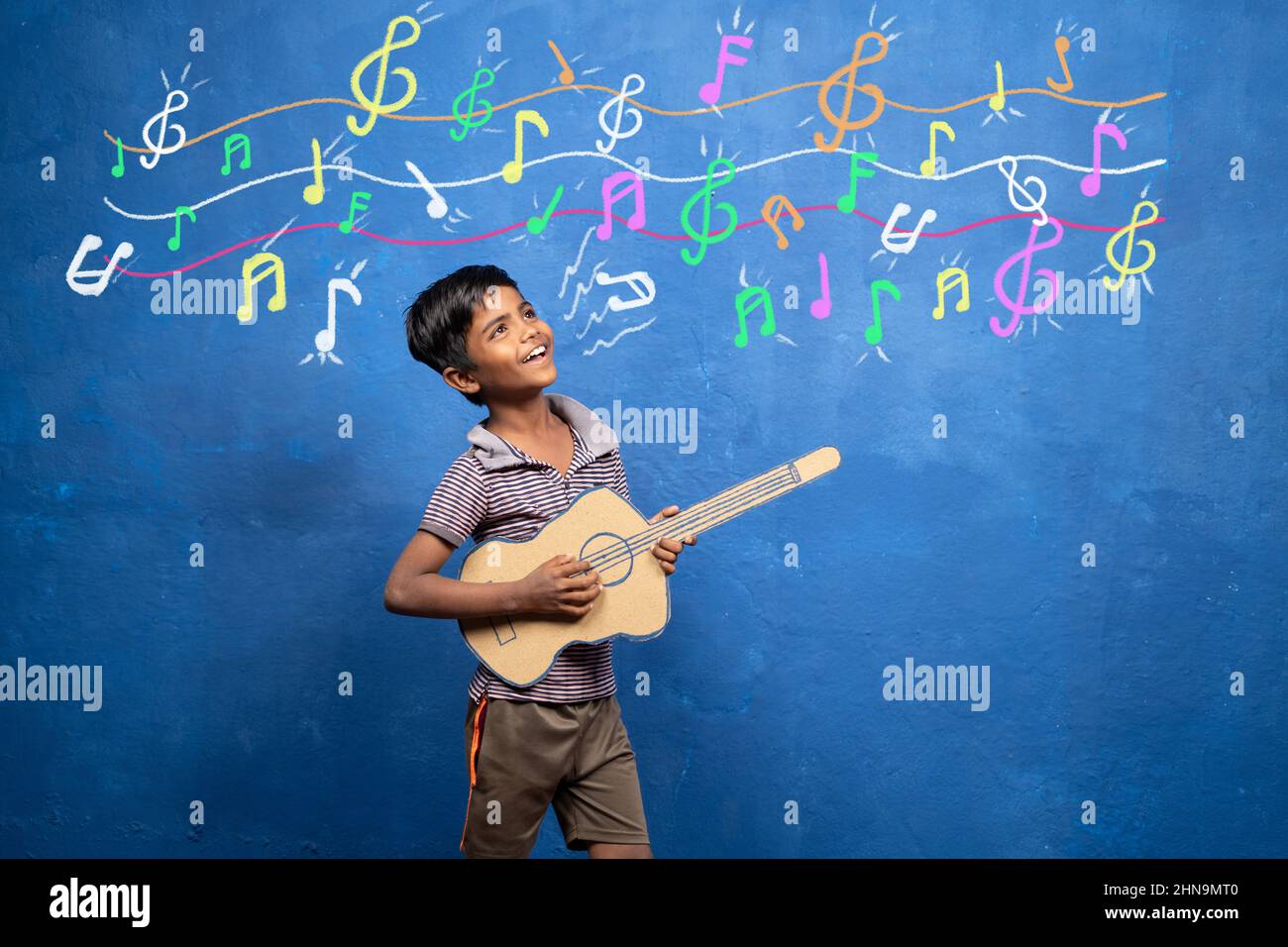 Happy Kid genießt es, mit Pappgitarre mit musikalischen Zeichen an der Wand zu spielen - Konzept der Unterhaltung, Armut, Kindheitstraum für Rockstar. Stockfoto