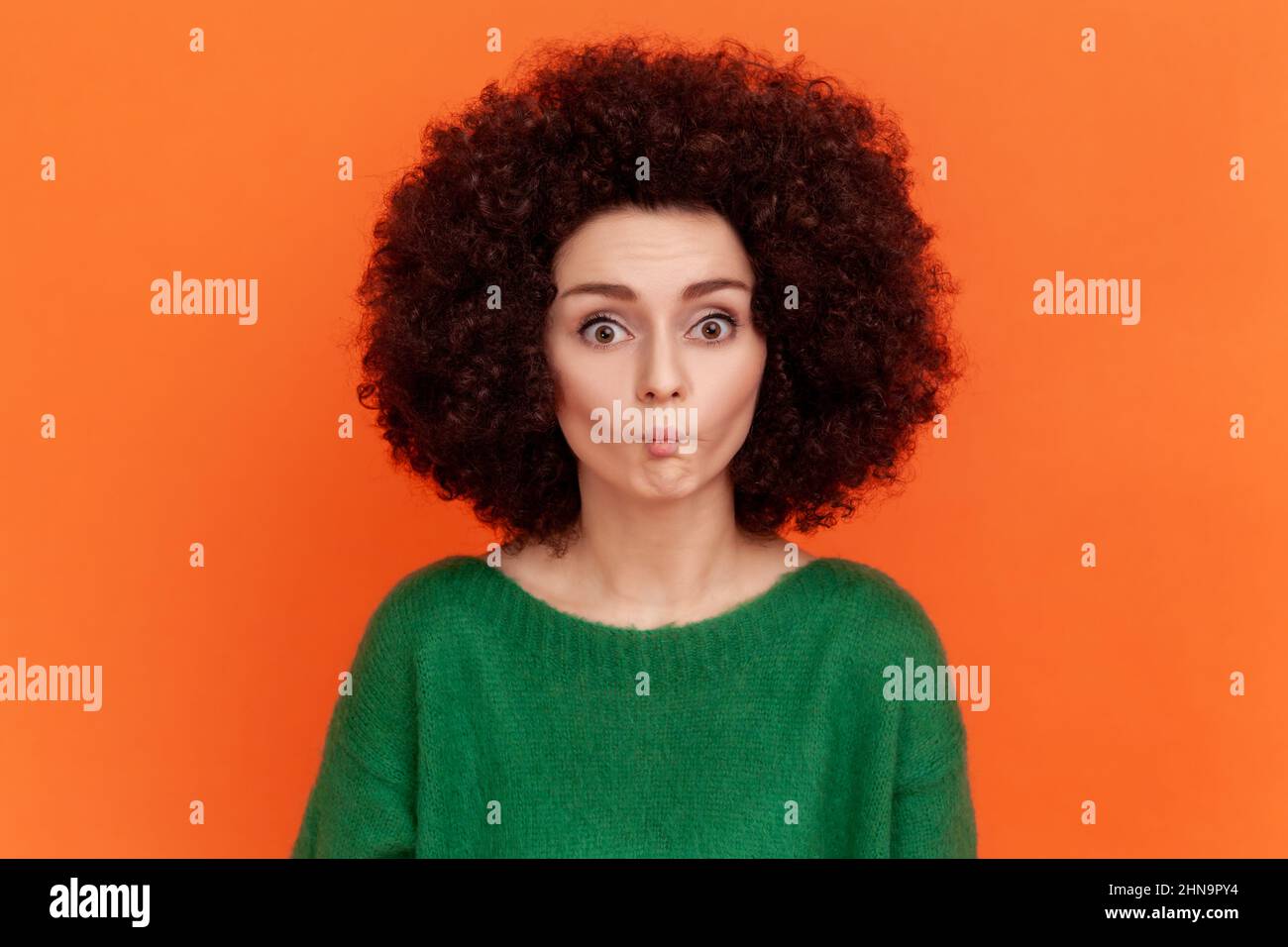 Lustige Frau mit Afro-Frisur in grünem Pullover macht Fisch Gesicht mit Puppen Lippen und sieht erstaunt, erstickend mit idiotisch komisch dummen Ausdruck. Innenaufnahme des Studios isoliert auf orangefarbenem Hintergrund. Stockfoto