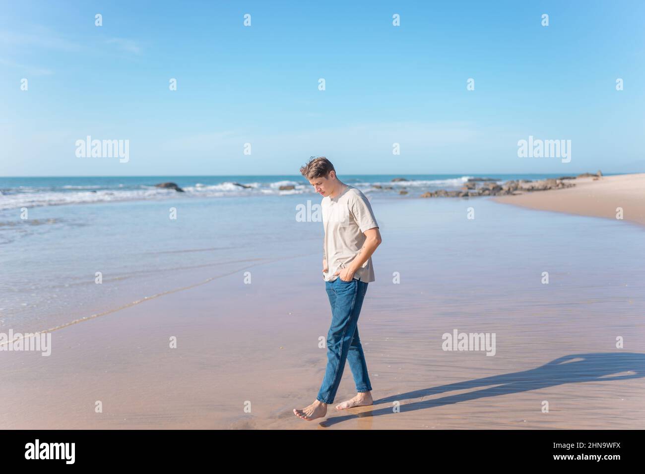 Attraktiver junger Kaukasier mit Vollarmtätowierung in legerer Kleidung, der barfuß am Strand läuft. Malerischer Blick aufs Meer, am Morgen. Hochwertige Fotos Stockfoto