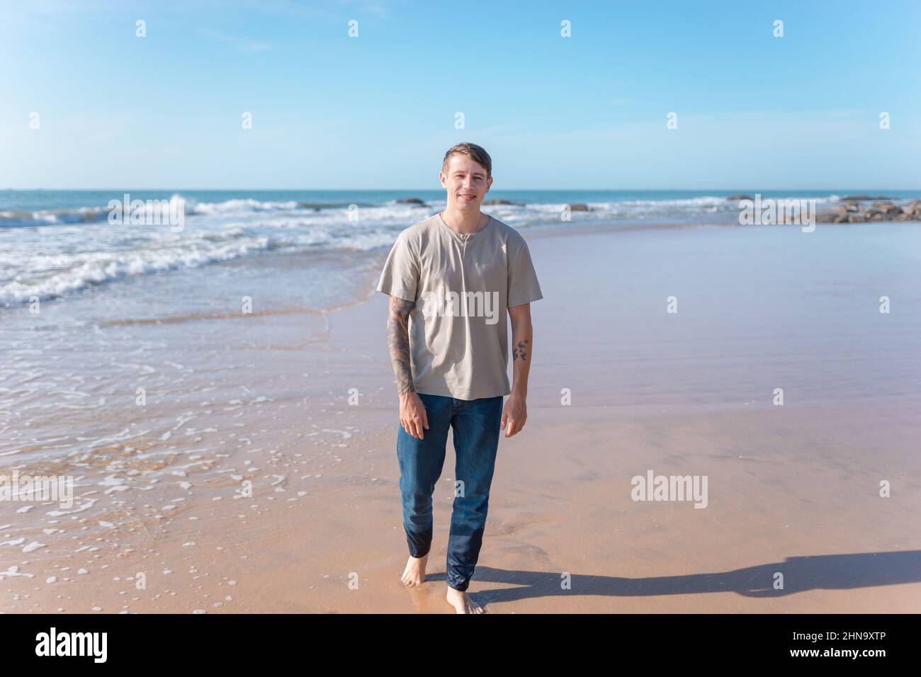 Attraktiver junger Kaukasier mit Vollarmtätowierung in legerer Kleidung, der barfuß am Strand läuft. Malerischer Blick aufs Meer, am Morgen. Hochwertige Fotos Stockfoto