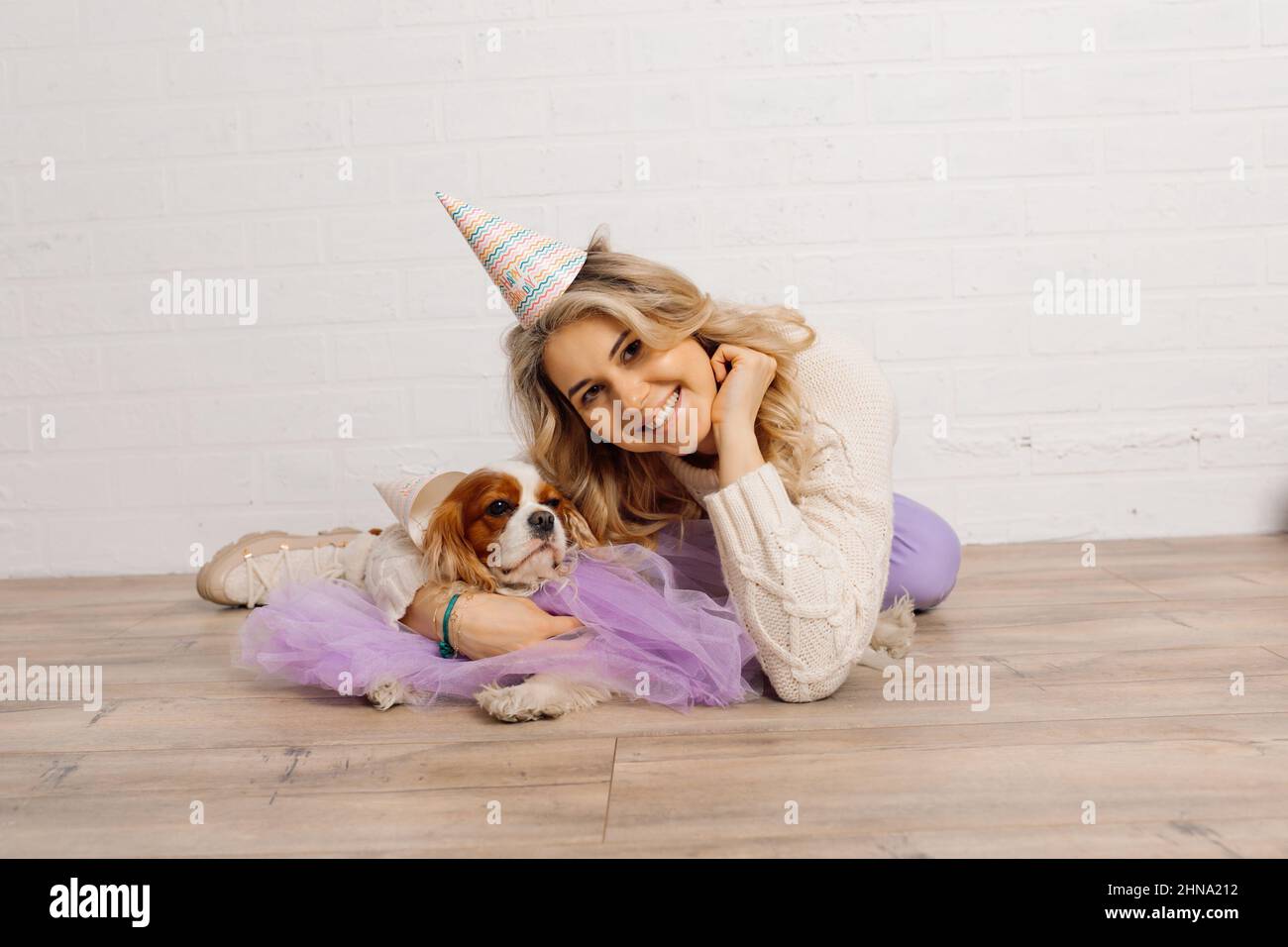 Die junge Blondine mit langen Haaren im Partyhut liegt auf dem Boden gegen die weiße Wand und umarmt ihren in Purpur gekleideten Hundekavalier King Charles Spaniel Stockfoto