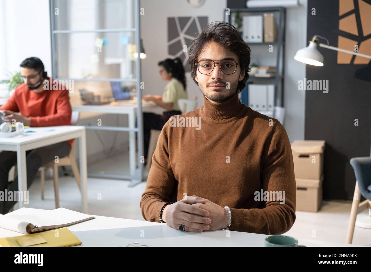 Porträt eines jungen Geschäftsmannes in einer Brille, der an seinem Arbeitsplatz sitzt und mit Kollegen im Hintergrund auf die Kamera blickt Stockfoto