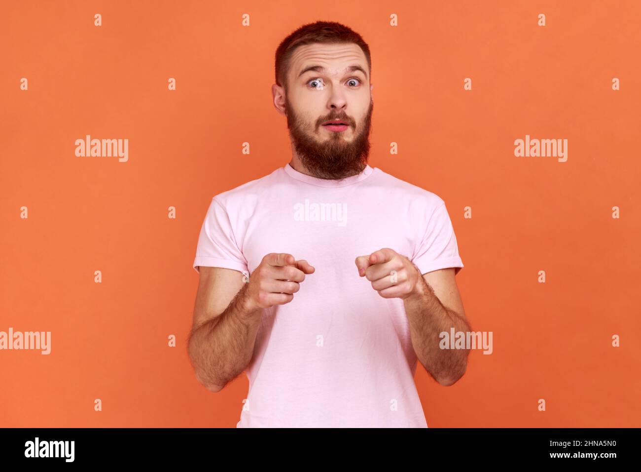 Porträt eines schockierten, überraschten bärtigen Mannes, der mit großen Augen und offenem Mund schaute, mit dem Finger auf die Kamera zeigte und dich verdächtigt, in einem rosa T-Shirt. Innenaufnahme des Studios isoliert auf orangefarbenem Hintergrund. Stockfoto