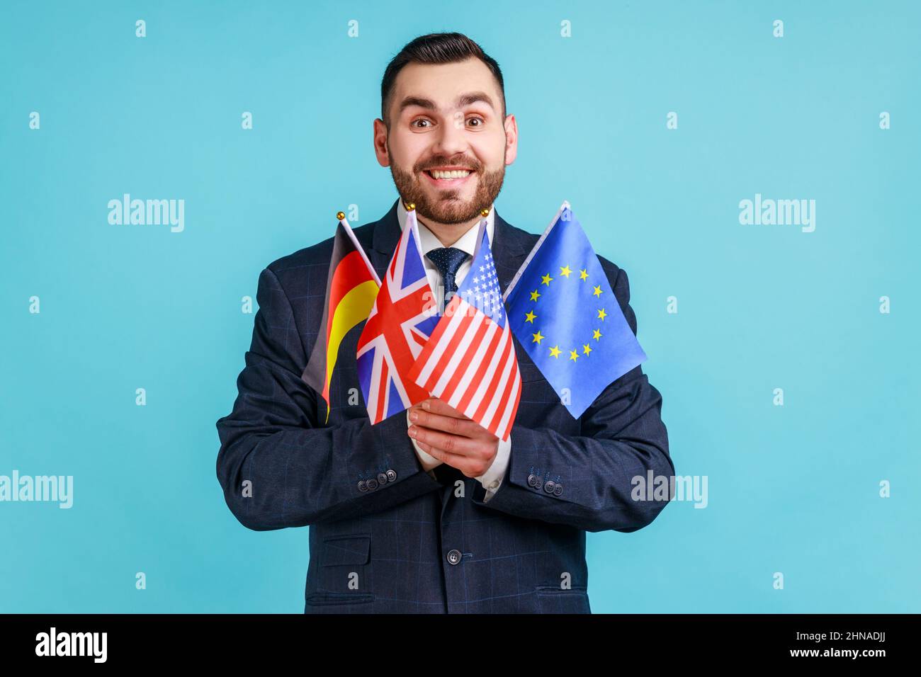 Positiver Mann mit Bart, der einen Anzug im offiziellen Stil trägt, der Flaggen verschiedener Länder hält, Sprachen lernt, die Kamera mit einem toothy Lächeln anschaut. Innenaufnahme des Studios isoliert auf blauem Hintergrund. Stockfoto