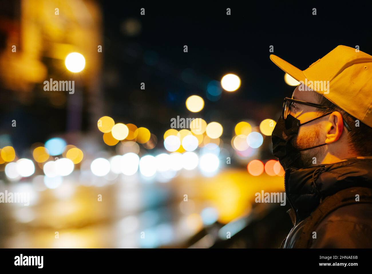 Porträt eines Mannes mit Brille und Gesichtsmaske vor Bokeh-Lichtern. Nachtaufnahme eines männlichen Fußgängers mit City Street Bokeh Hintergrund Stockfoto