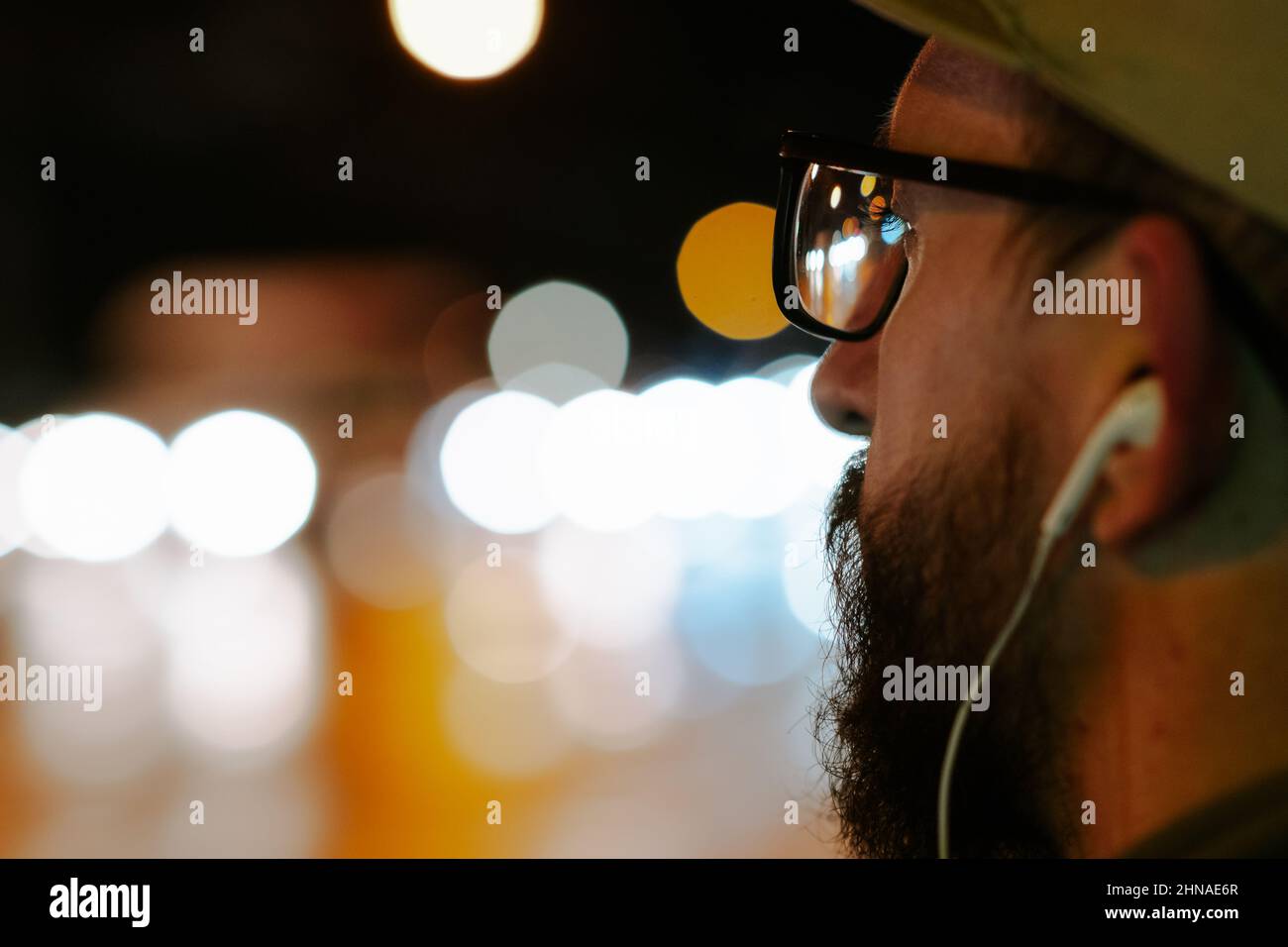 Nahaufnahme eines Mannes mit Brille und Ohrhörer vor Bokeh-Lichtern in der Nacht. Männlicher Fußgänger mit Stadtstraße Bokeh Hintergrund, flach Stockfoto