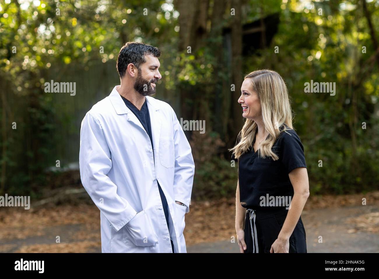 Ein Arzt mit dunklem Haar und einem Bart in schwarzen Peelings und einem weißen Laborkittel, der draußen steht und mit einer medizinischen Fachkrankenschwester spricht Stockfoto