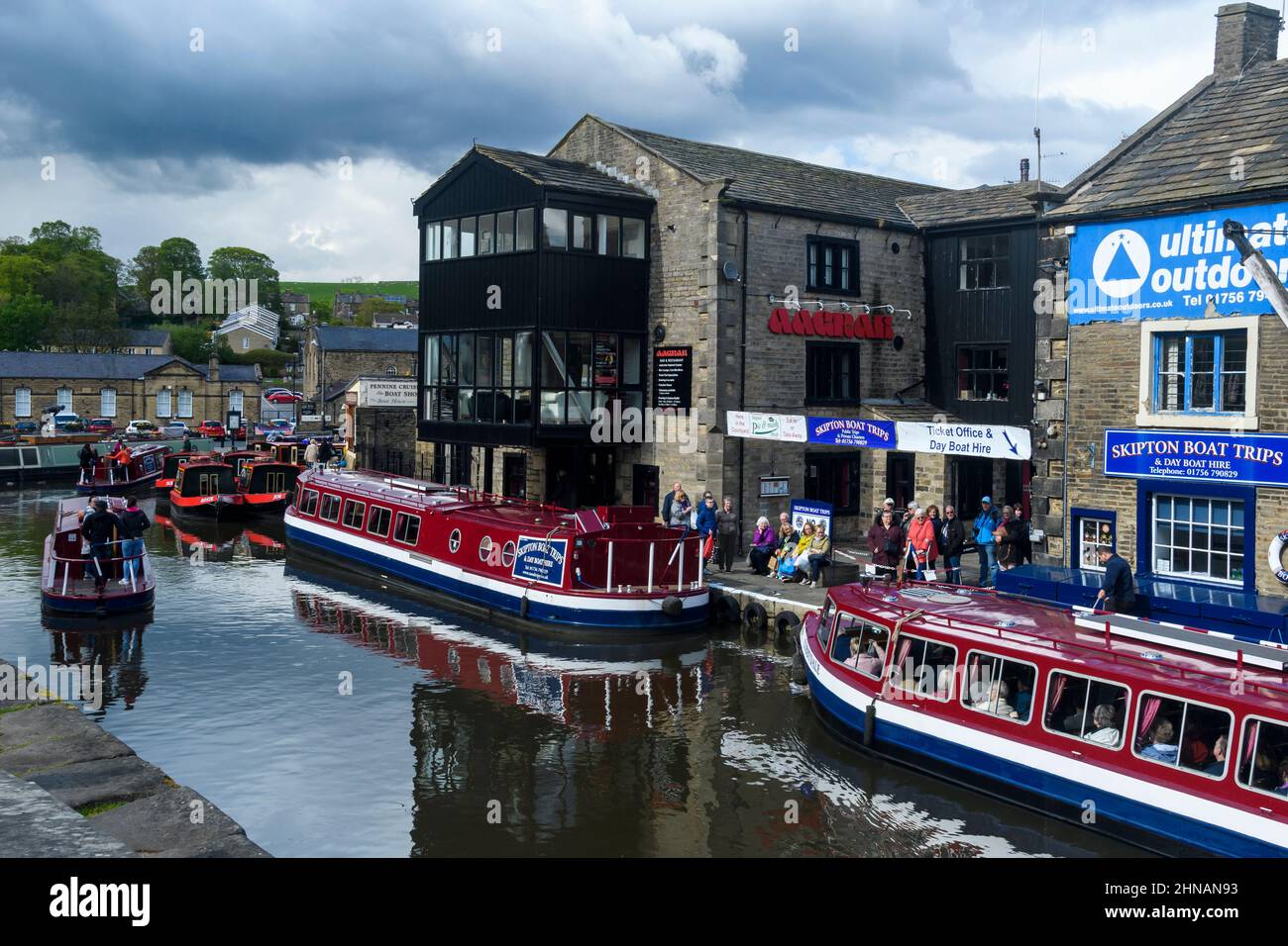 Geschäftiges, beliebtes Touristenfreizeiterlebnis auf dem Wasser (rote Bootsanlegestellen, Männer, Frauen, die Schlange stehen) - landschaftlich reizvoller Leeds-Liverpool Canal, Yorkshire, England, Großbritannien. Stockfoto