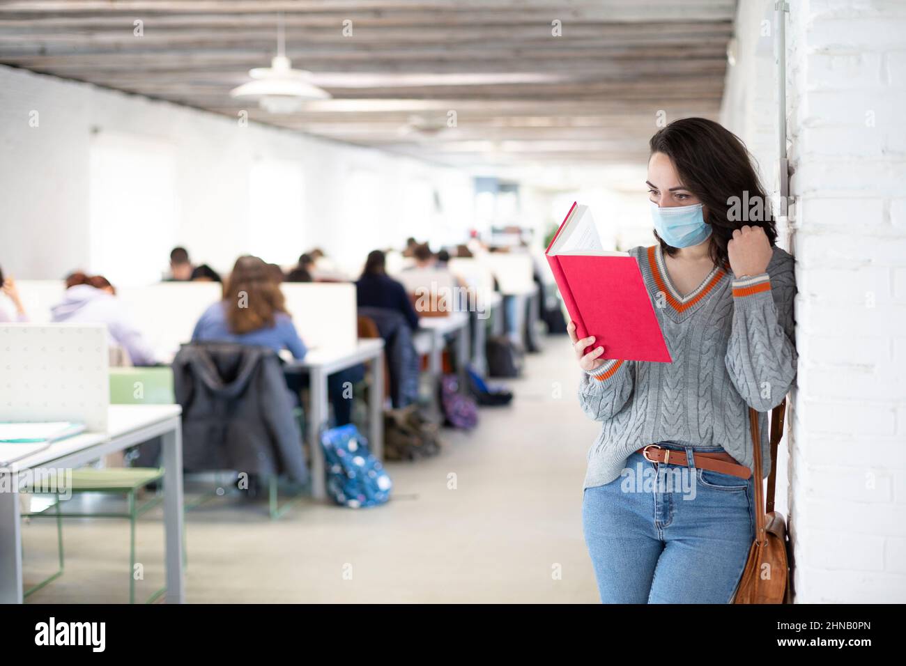Junge Schülerin mit Gesichtsmaske beim Lesen eines Buches in einem Klassenzimmer. Präsenzunterricht an der Universität. Platz für Text. Stockfoto