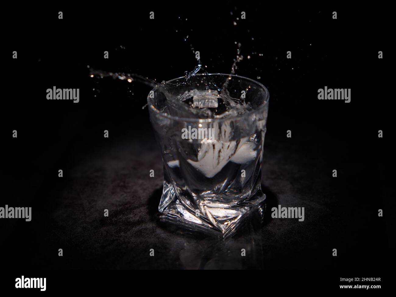 Wasserspritzer aus einem Eisstück, das in ein transparentes Glas mit schwarzem Hintergrund fällt Stockfoto