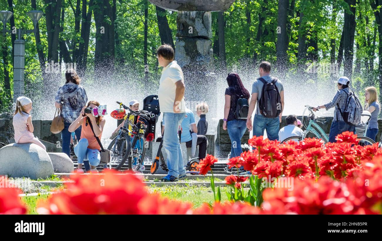 An einem sonnigen, heißen Tag im Stadtpark entspannen sich die Menschen mit ihren Familien in der Nähe des Brunnens. Eltern mit Kindern werden fotografiert. Wasser spritzt Stockfoto