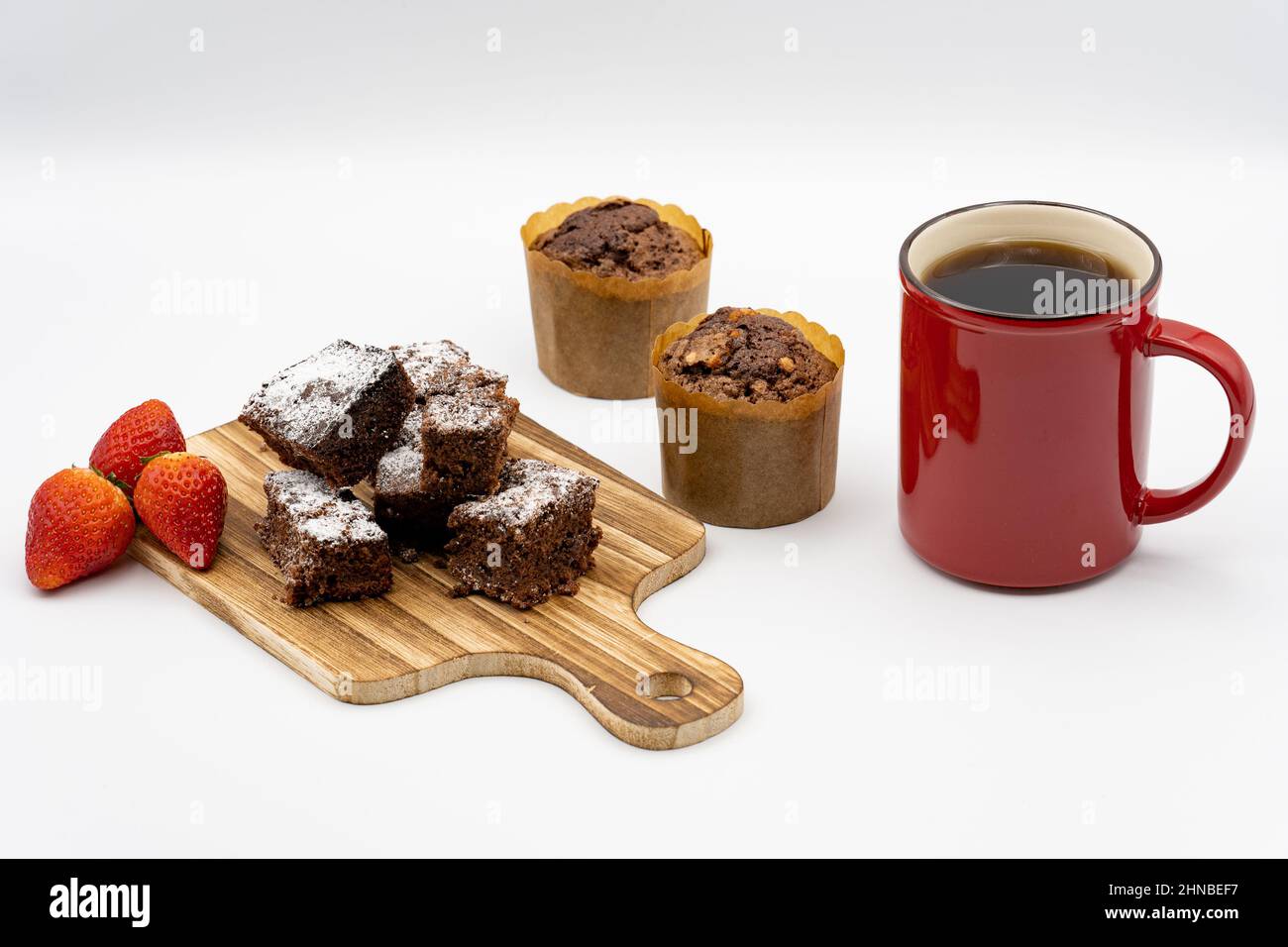 Brownies auf Holzbrett, Schokolade-Muffins, verschiedene Pralinen auf einem weißen Teller, rote Kaffeetasse, Erdbeeren und weißer Hintergrund Stockfoto
