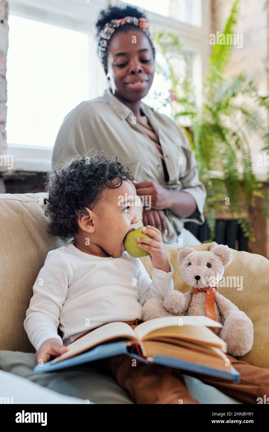 Süßes afroamerikanisches Kleinkind mit lockigem Haar, das mit Buch und Spielzeugbär im Sessel sitzt und unter Kontrolle der Mutter Apfel isst Stockfoto