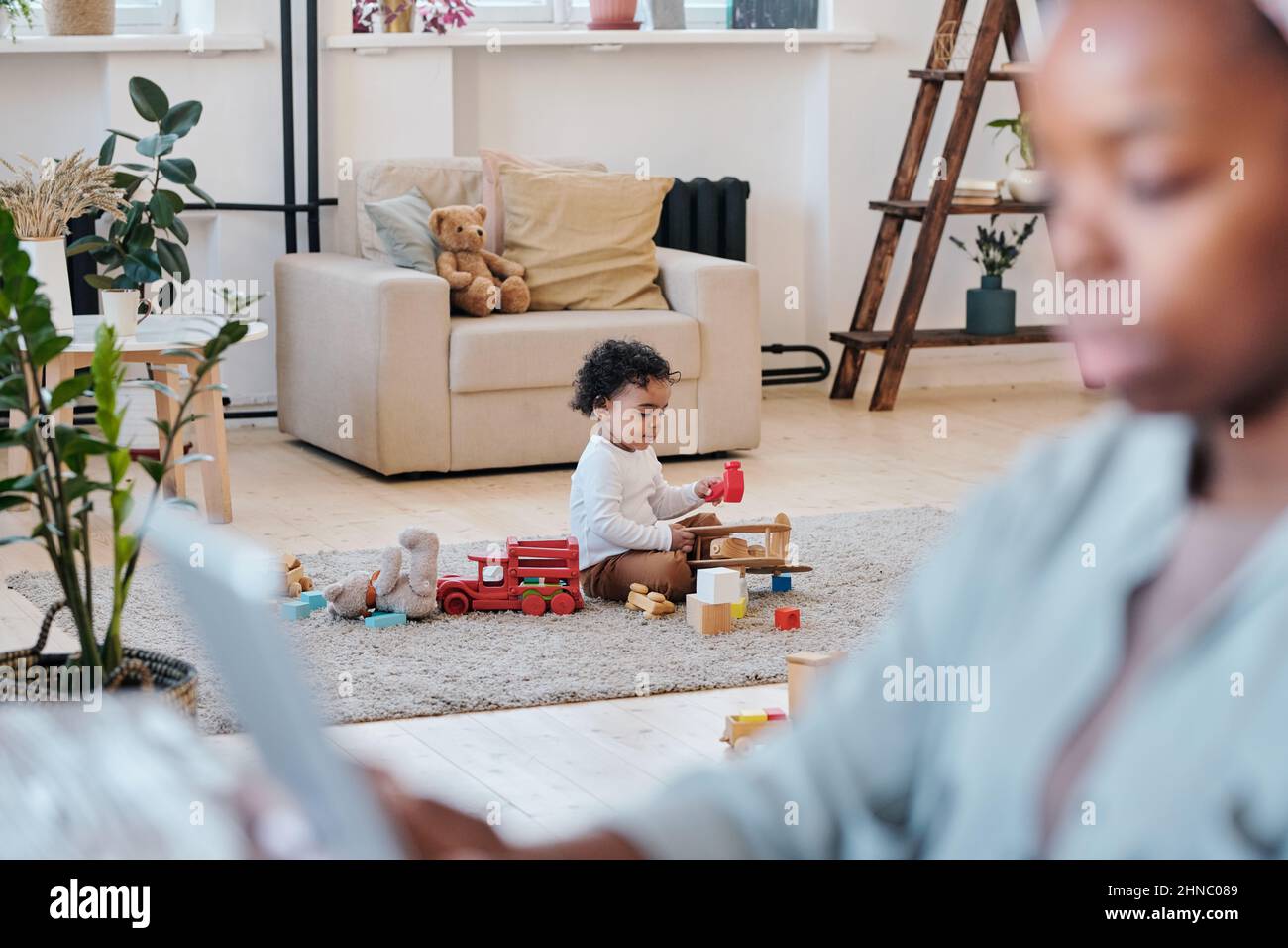 Afroamerikanisches Kleinkind sitzt auf dem Teppich und spielt allein mit Spielzeug, während seine Mutter arbeitet, konzentriert sich auf den Hintergrund Stockfoto