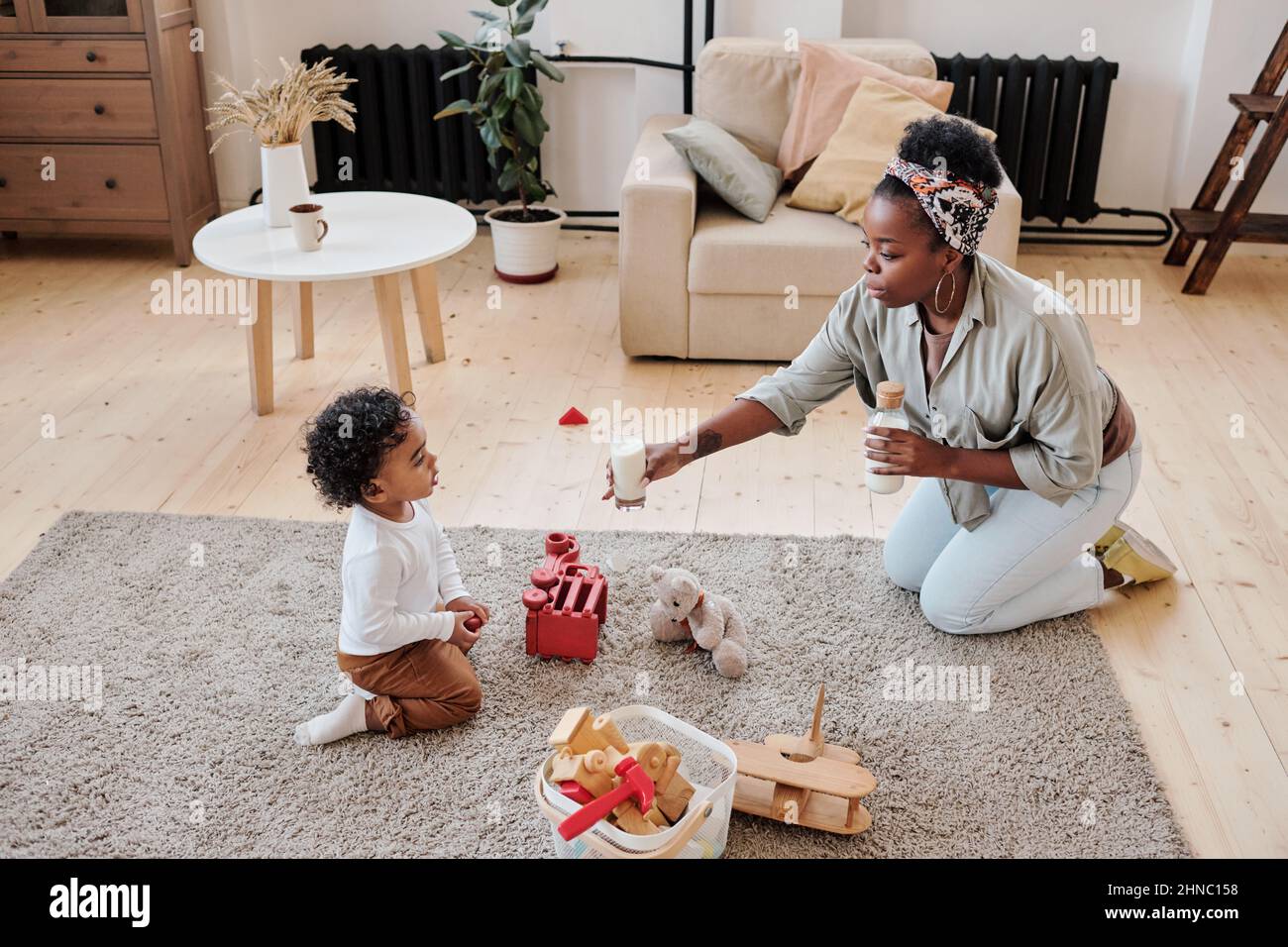 Vorsichtige junge afroamerikanische Mutter im Kopftuch, die auf dem Boden sitzt und dem kleinen Sohn ein Glas Milch gibt Stockfoto