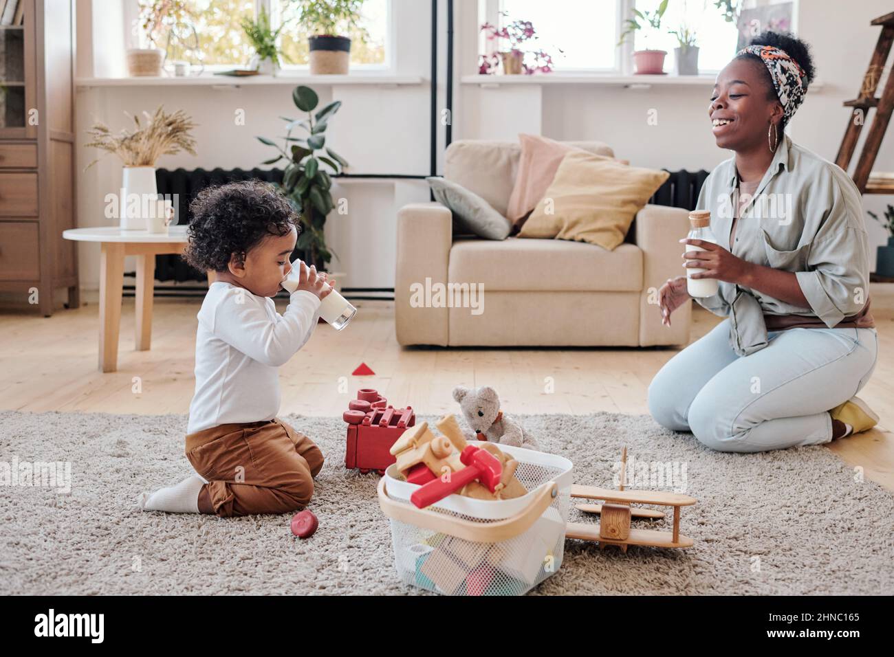 Kleiner afroamerikanischer Junge, der mit Spielzeug auf dem Teppich sitzt und Milch trinkt, während eine positive junge Mutter ihn anschaut Stockfoto