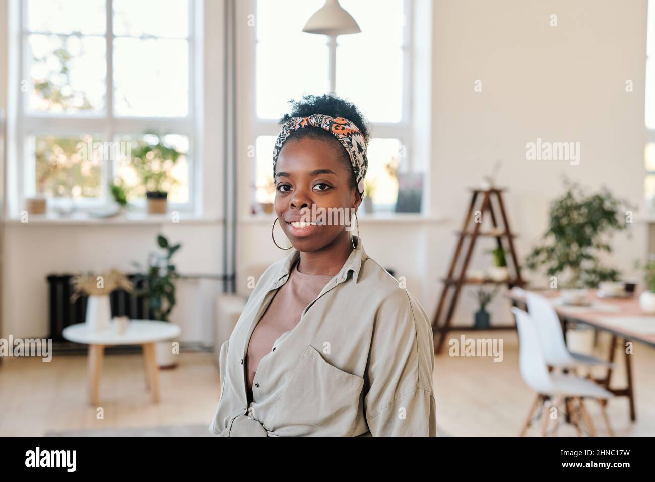 Porträt einer lächelnden jungen afroamerikanischen Frau mit Kopftuch und runden Ohrringen, die im modernen Coworking Space stehen Stockfoto