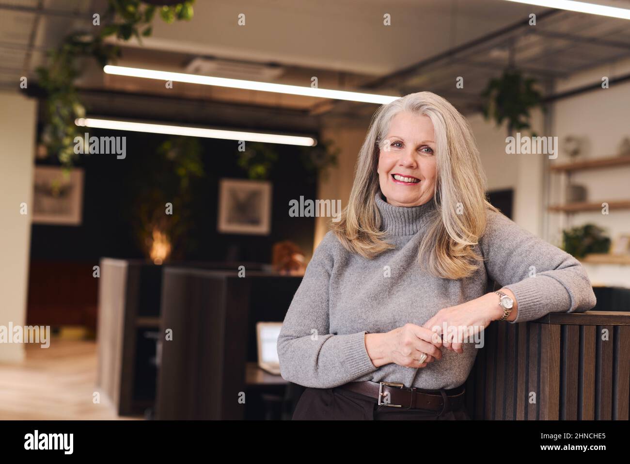 Porträt einer selbstbewussten älteren weißen Frau mit langen grauen Haaren, die lächelt und die Kamera im Coworking Space anschaut Stockfoto