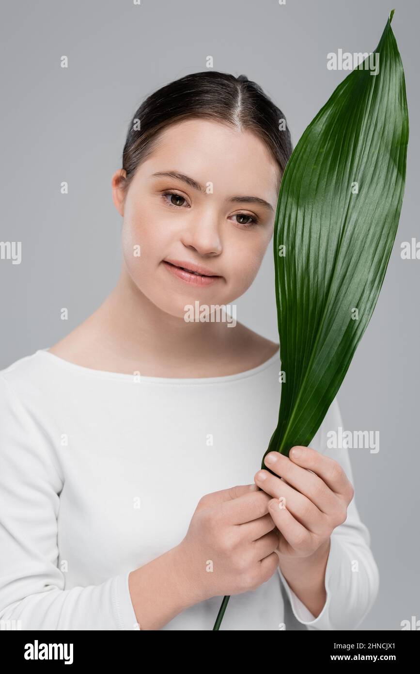 Lächelnde Brünette Frau mit Down-Syndrom hält Blatt isoliert auf grau Stockfoto