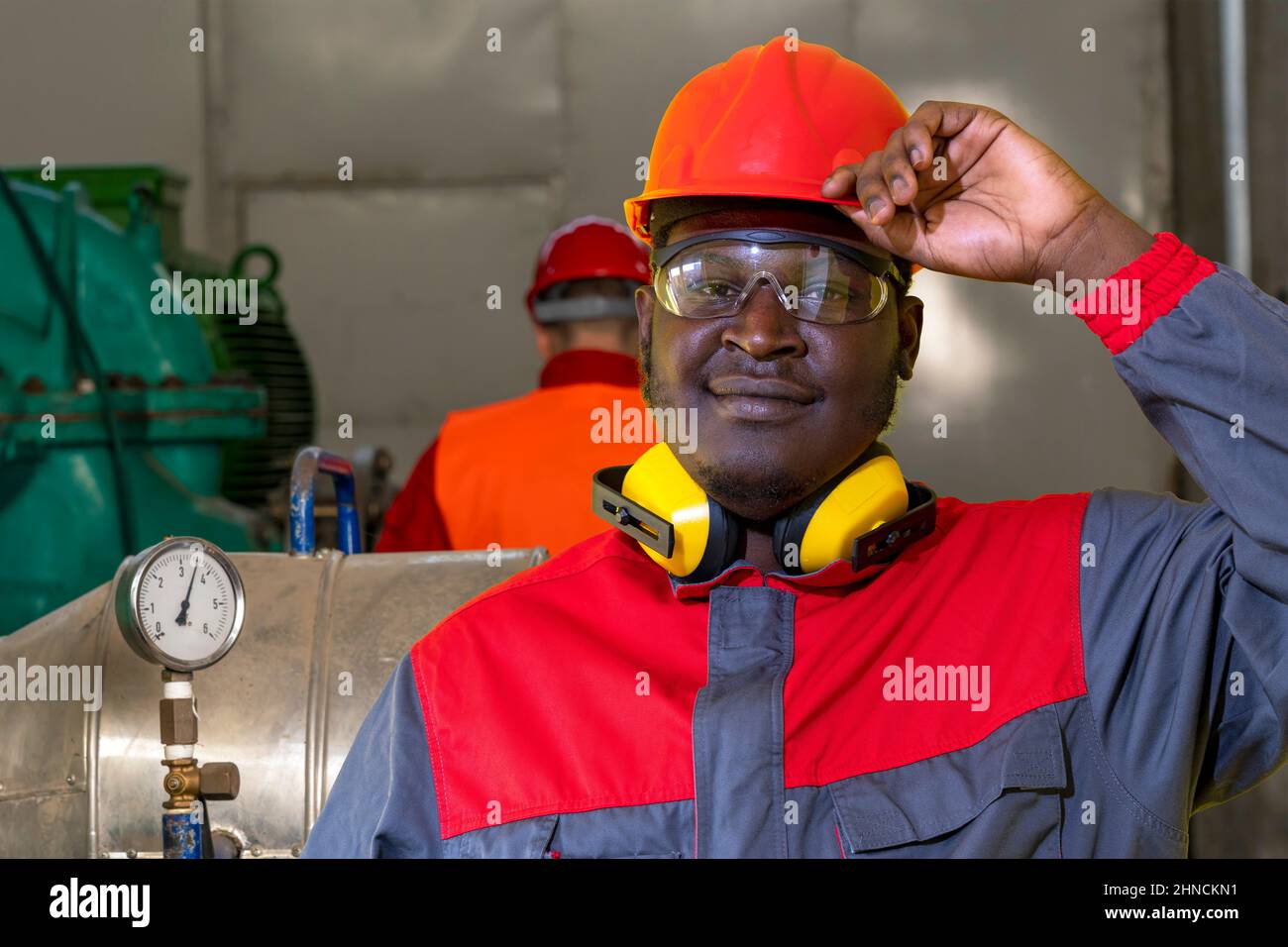 Schwarzer Industriearbeiter Mit Rotem Helm, Schutzbrille, Gehörschutzausrüstung Und Arbeitskleidung. Stockfoto