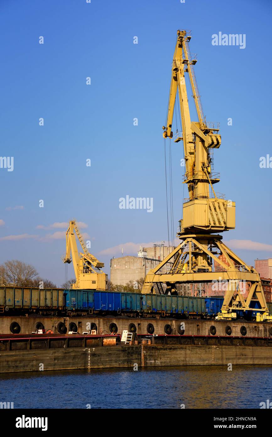 Am sonnigen Tag lädt ein Kran aus dem Flusshafen die oben offenen Gondelautos. Leere Flussschiffe oder Lastkähne, die am Pier vertäut sind, leere Autos, die zum Laden bereit sind. Stockfoto