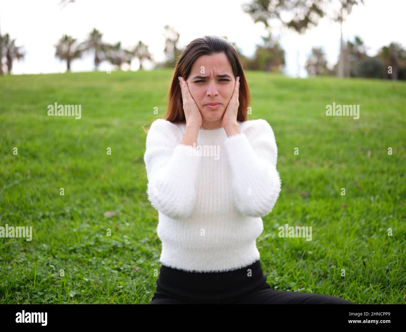 Eine junge Frau im Park, die auf dem Gras sitzt und ihre Ohren wegen eines sehr lauten Lärms bedeckt. Stockfoto