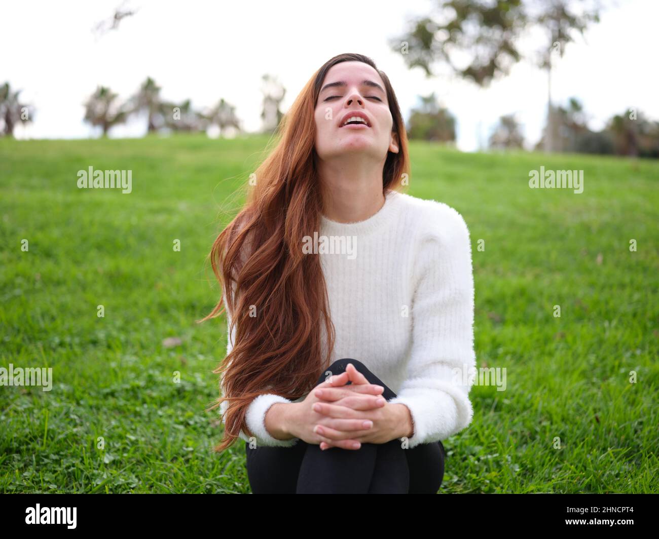 Eine entspannte junge Frau im Park sitzt mit geschlossenen Augen auf dem Gras und atmet frische Luft Stockfoto