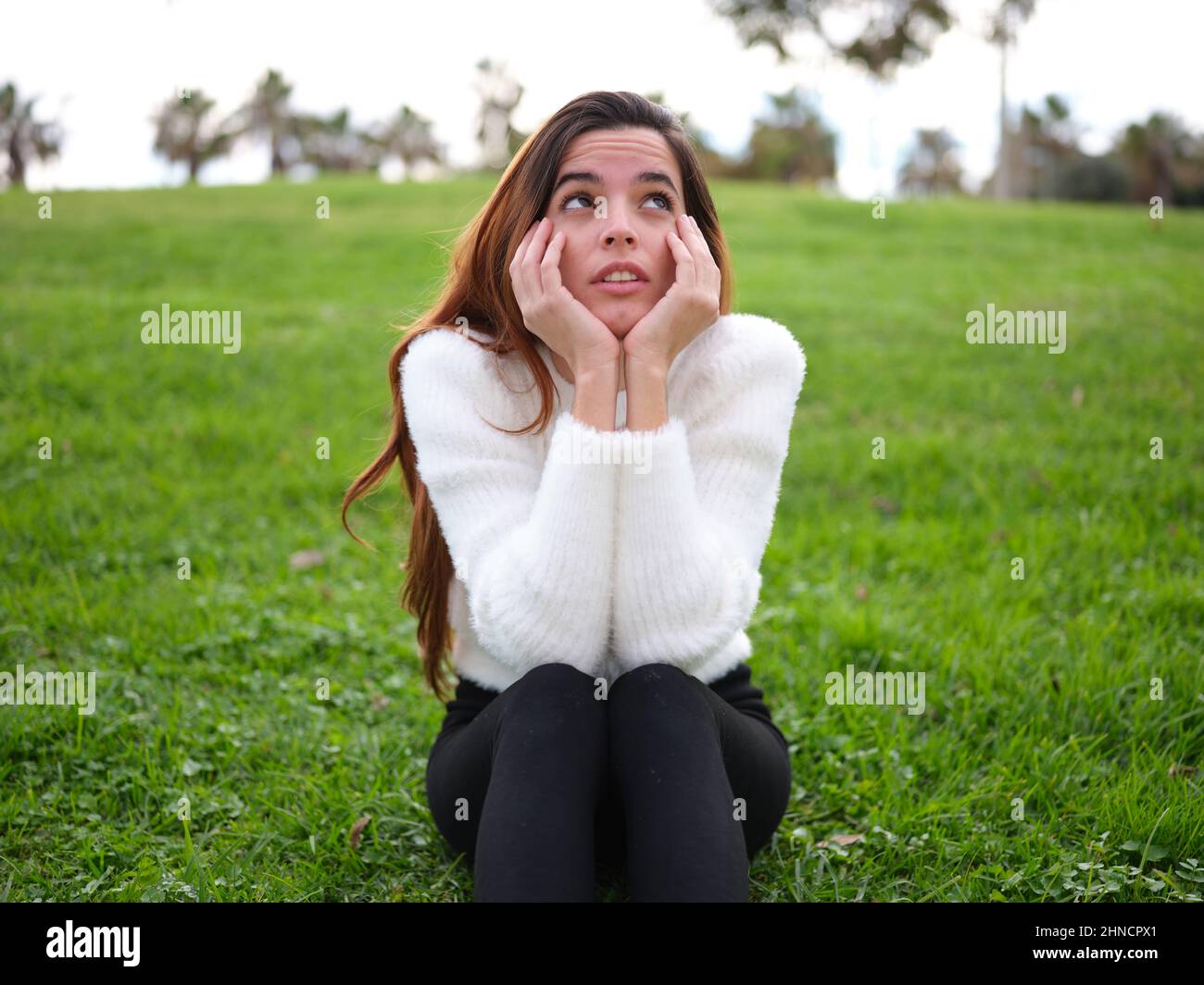 Eine junge Frau im Park, die auf dem Gras sitzt und mit ihren Händen auf ihrem Kinn aufschaut, sich vorstellt oder denkt. Stockfoto