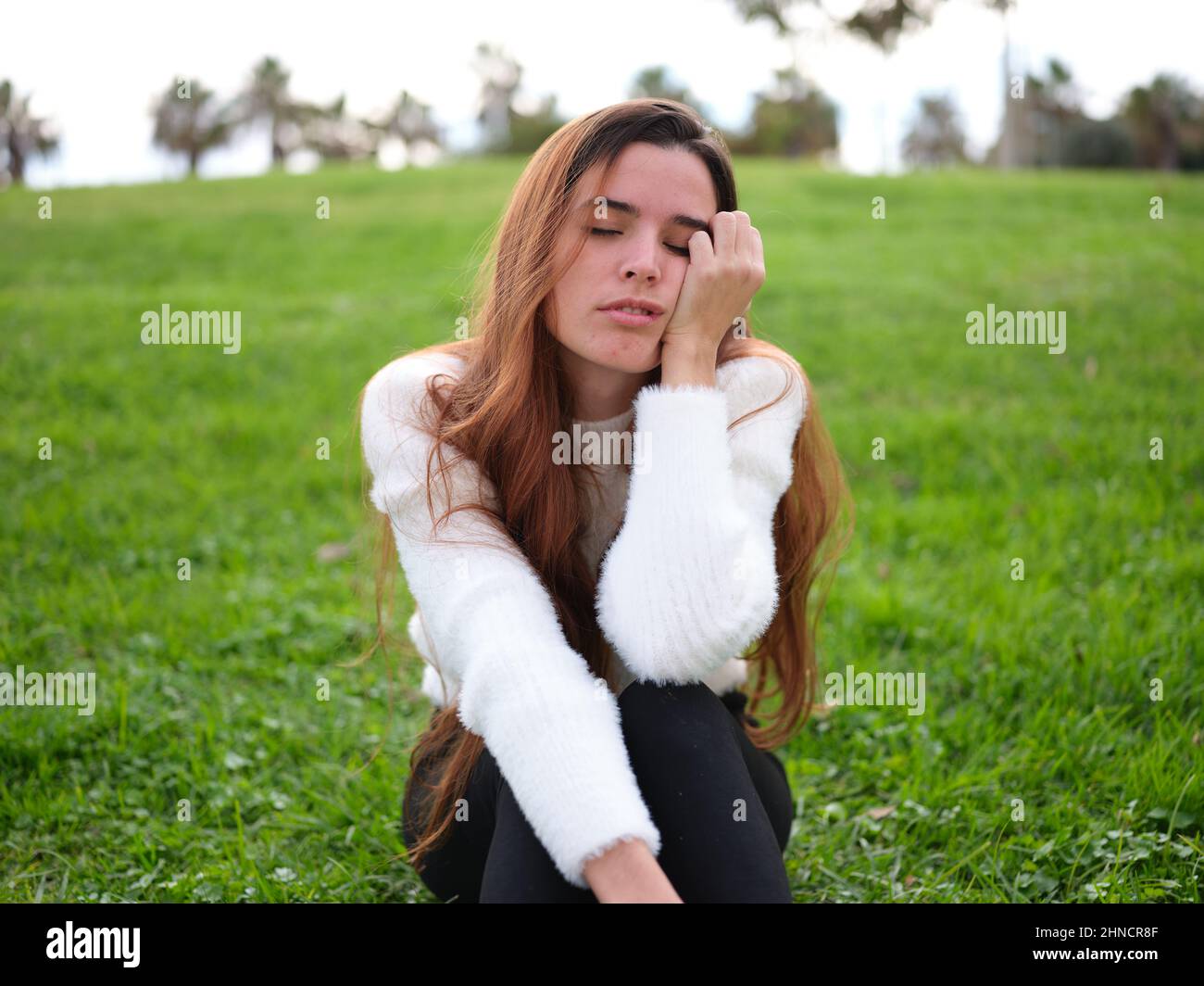 Eine junge Frau im Park sitzt auf dem Gras, mit einer Hand auf ihrem Gesicht und schläft ein. Stockfoto