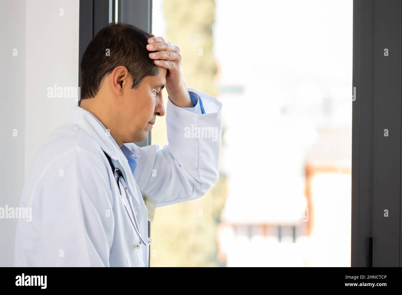 Aufnahme eines jungen männlichen Arztes, der gestresst aussicht, während er an einem Fenster in einem Krankenhaus steht Stockfoto