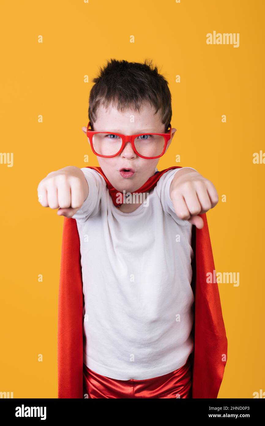 Isolierter Junge in Brille und rotem Superhelden-Kostüm, der die Kamera anschaut, während er vorgibt, im Studio auf gelbem Hintergrund zu fliegen Stockfoto