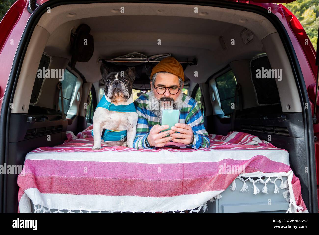 Konzentrierter reifer Rüde, der während einer Fahrt auf dem Land im Mini-Camper mit der französischen Bulldog auf dem Smartphone herumliegt Stockfoto