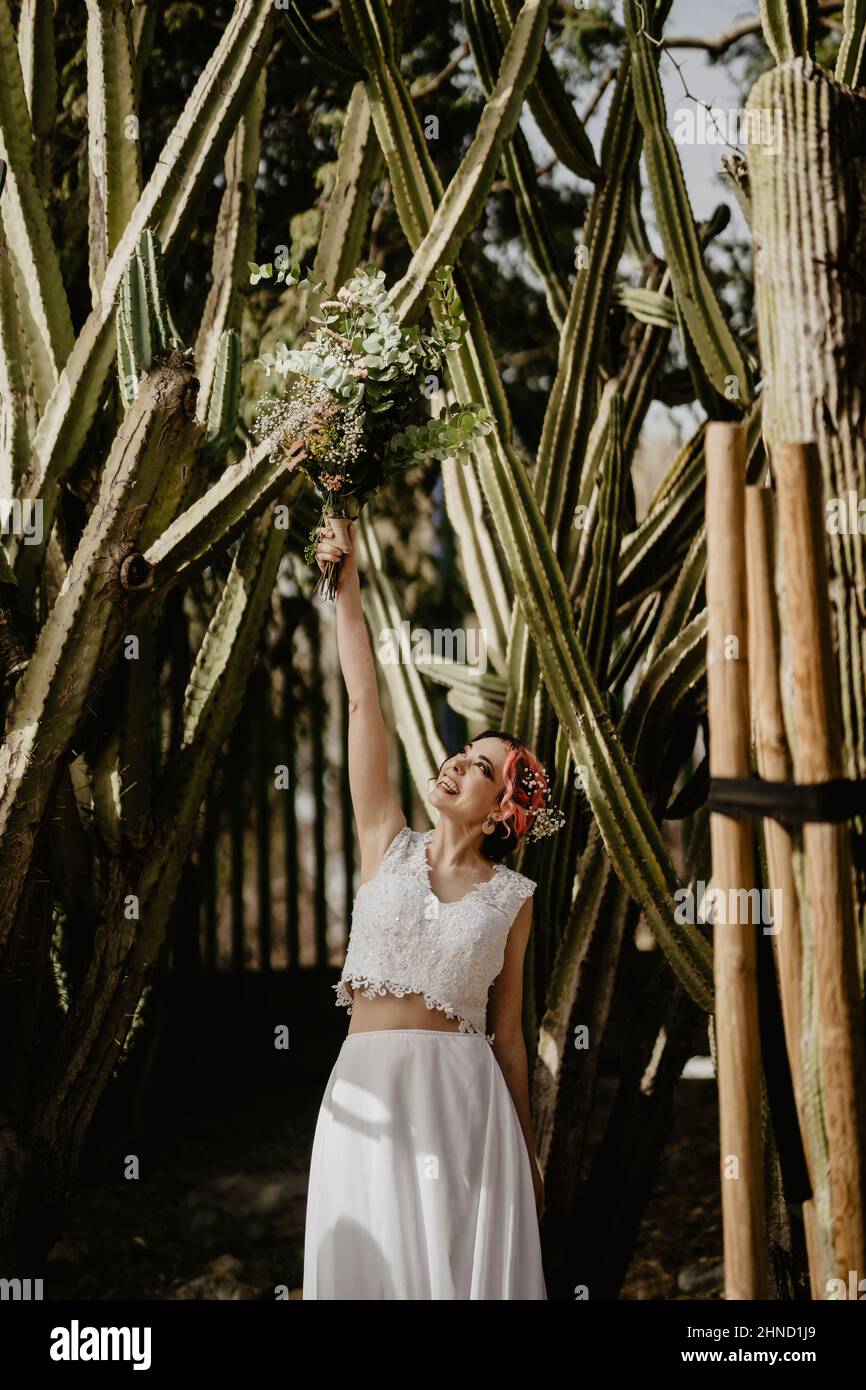 Strahlende junge Frau in weißem Oberteil und Rock lächelt und hebt den Arm mit Brautstrauß, während sie am Hochzeitstag im Garten in der Nähe großer Kakteen steht Stockfoto