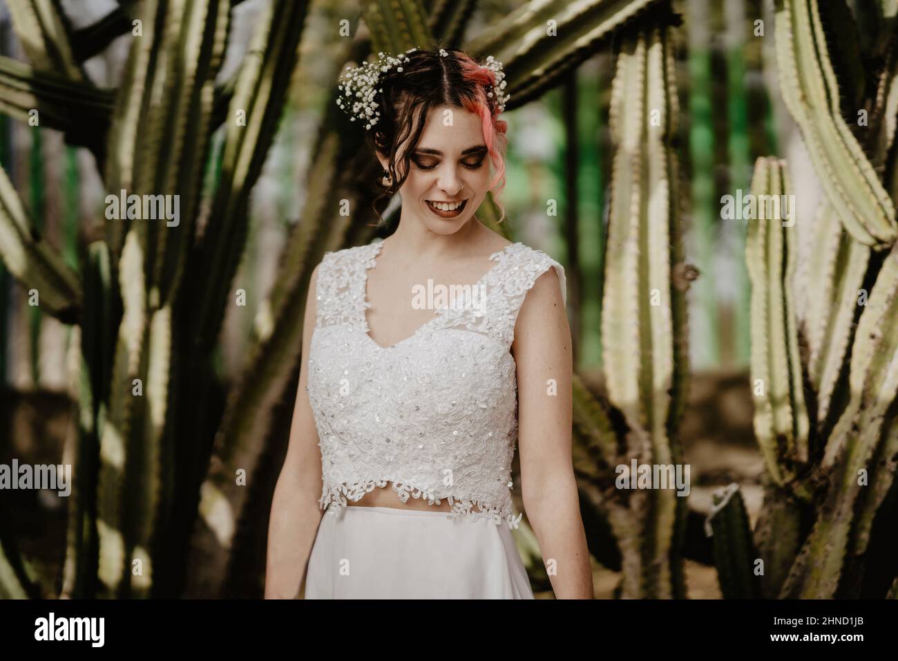 Strahlende junge Frau in weißem Oberteil und Rock lächelt, während sie am Hochzeitstag in der Nähe großer Kakteen im Garten steht und nach unten blickt Stockfoto