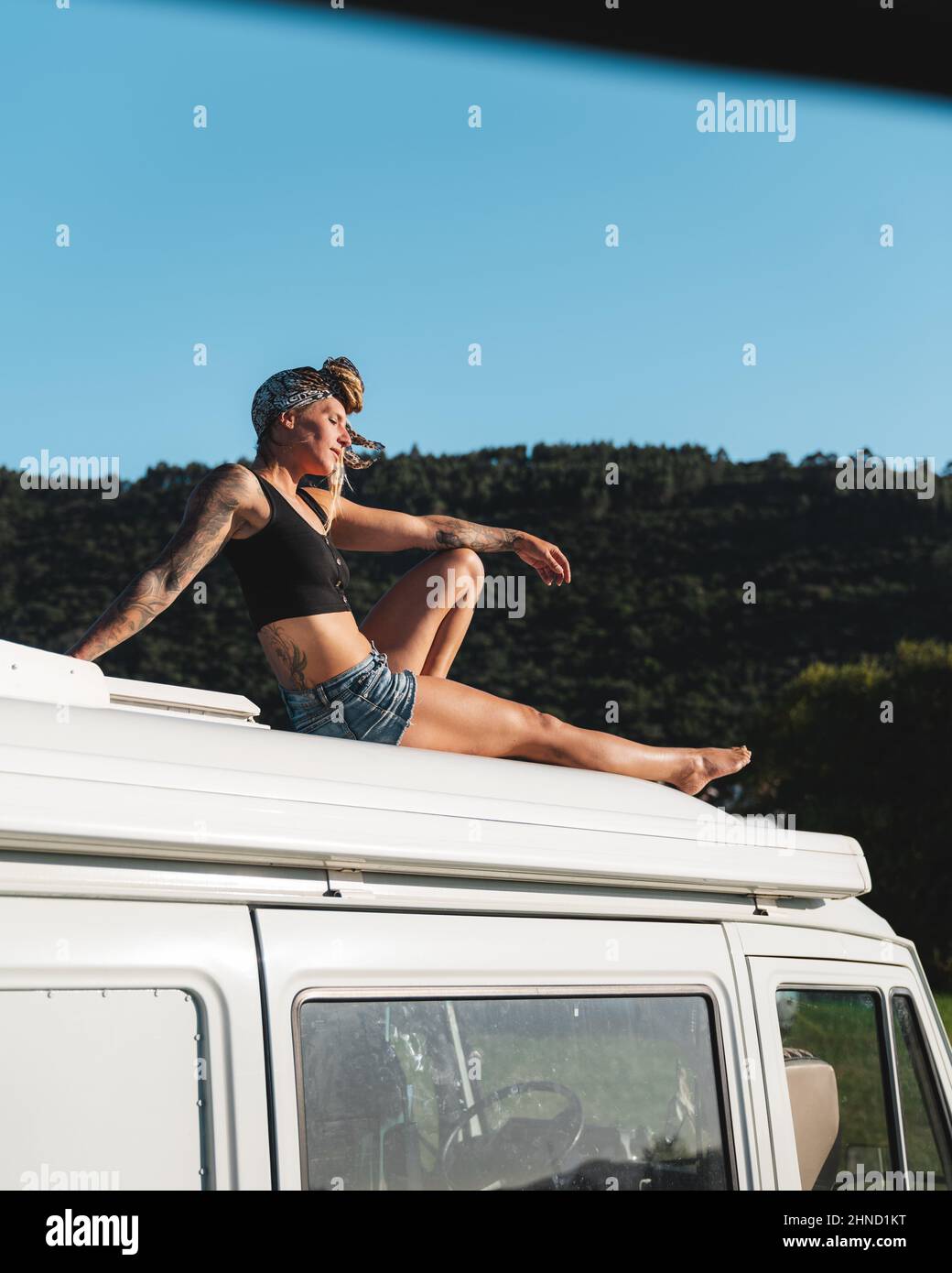 Ganzkörper-Seitenansicht der angenehmen barfuß nachdenklichen Hippie weibliche Reisende sitzen mit geschlossenen Augen auf dem Dach des weißen Camping van in der Natur geparkt d Stockfoto