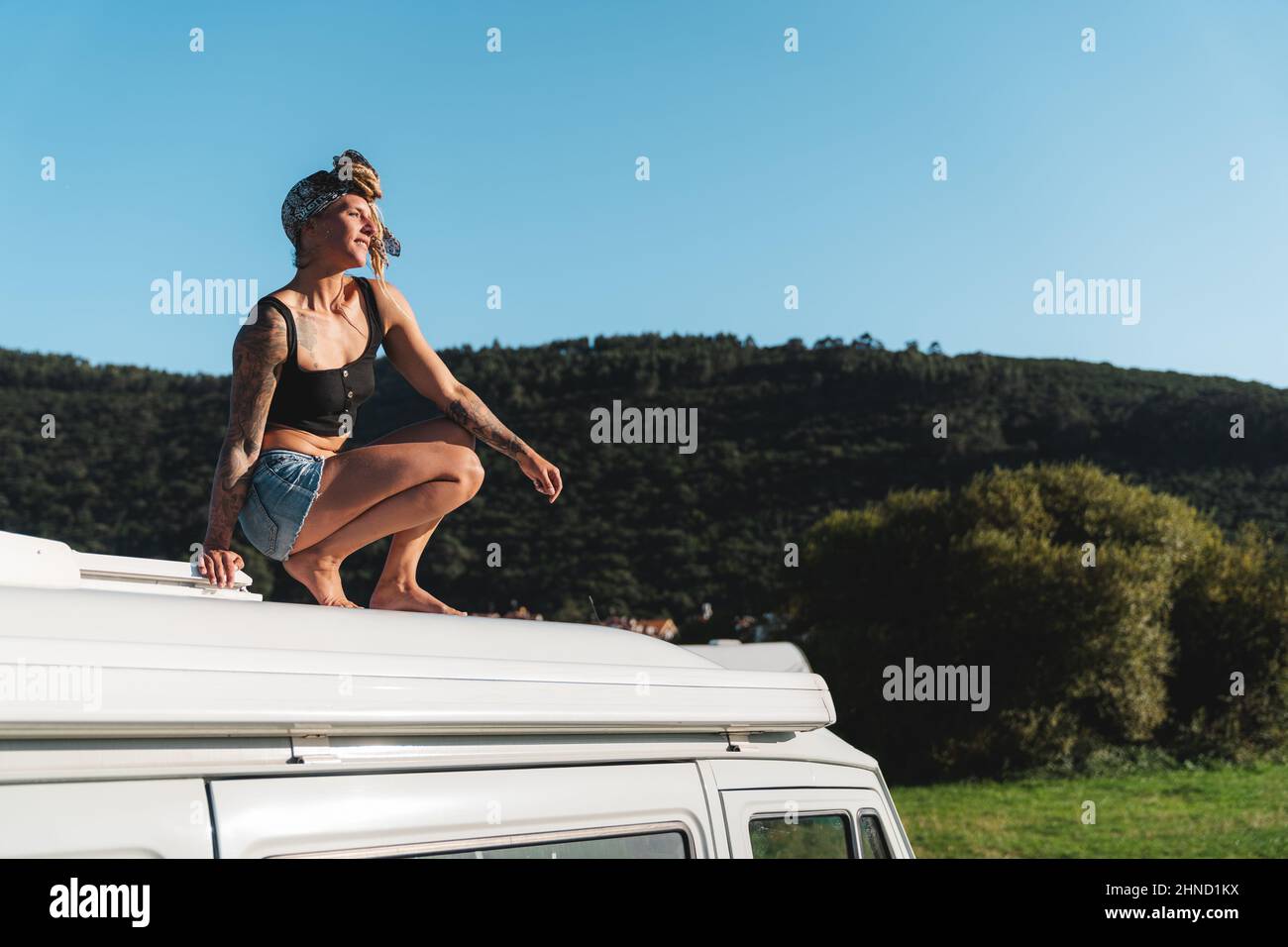 SID Blick auf den ganzen Körper von angenehmen barfuß nachdenklich Hippie weibliche Reisende sitzen auf dem Dach des weißen Camping van in der Natur während der Fahrt geparkt Stockfoto