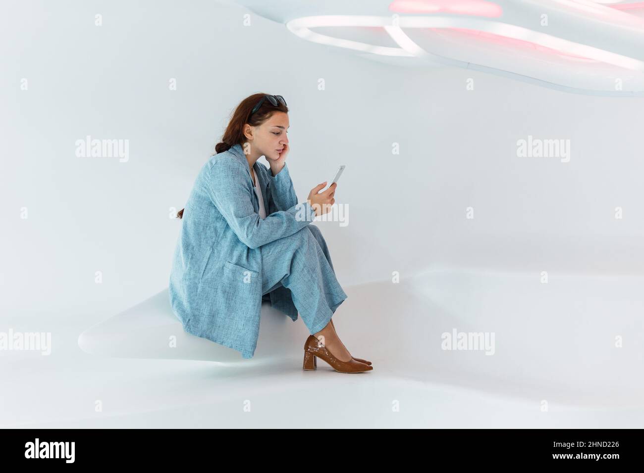 Seitenansicht des gesamten Inhalts Frau in eleganter Kleidung scrollende soziale Medien auf dem Handy, während sie in einem hellen modernen Raum mit weißer Wand sitzt Stockfoto