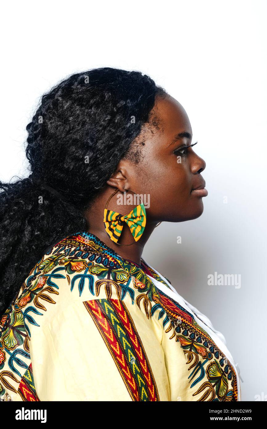 Seitenansicht einer jungen kurvigen Frau mit Afro-Frisur, die ein farbenfrohes afrikanisches Hemd und Ohrringe trägt. Sie schaut weg, während sie auf weißem Wandhinterst steht Stockfoto