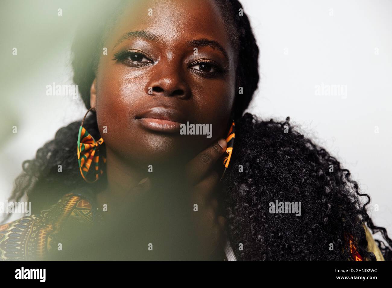 Porträt einer jungen kurvigen Frau in stilvoller afrikanischer Kleidung, die im hellen Raum auf die Kamera schaut. Sie hat unfokussierte Pflanzen im Foregrou Stockfoto