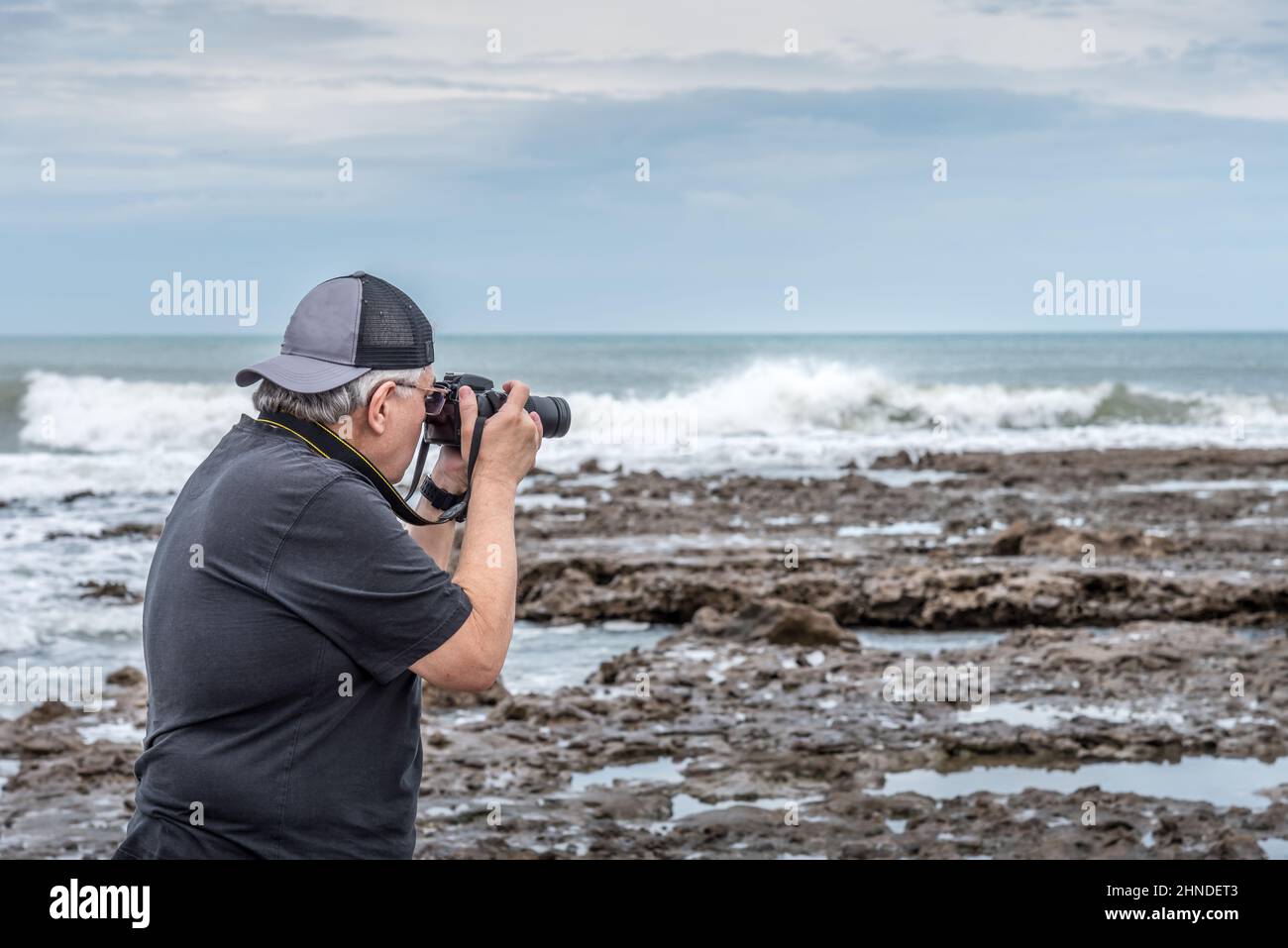 Halbkörper-Rückansicht eines erwachsenen Mannes mit Kappe, die ein Foto auf den Felsen in der Nähe des Meeres macht. Stockfoto