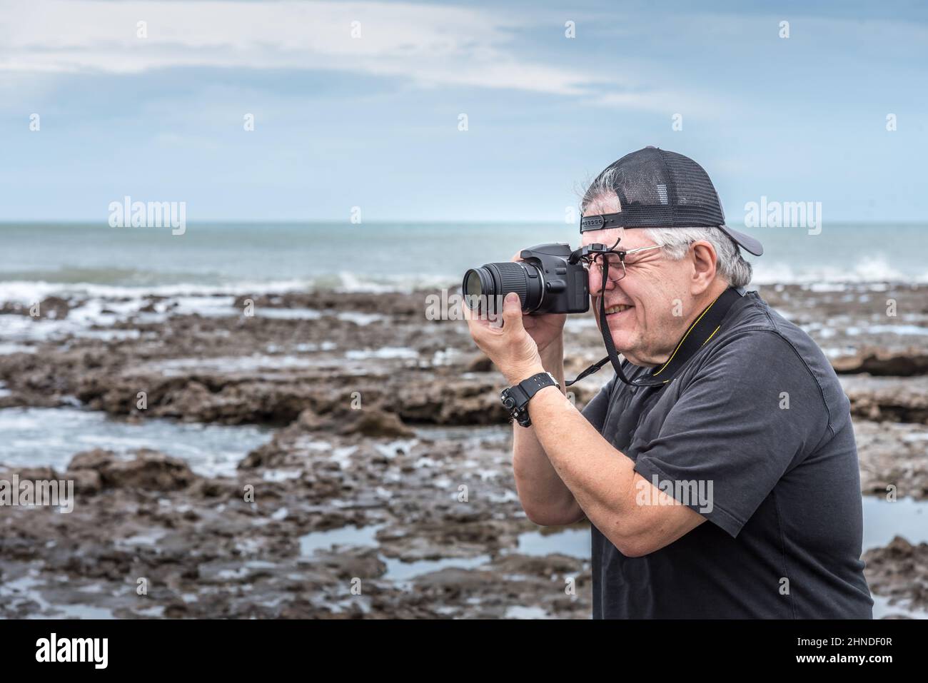 Halbkörperansicht eines erwachsenen Mannes mit Kappe, die ein Foto auf den Felsen in der Nähe des Meeres macht. Stockfoto