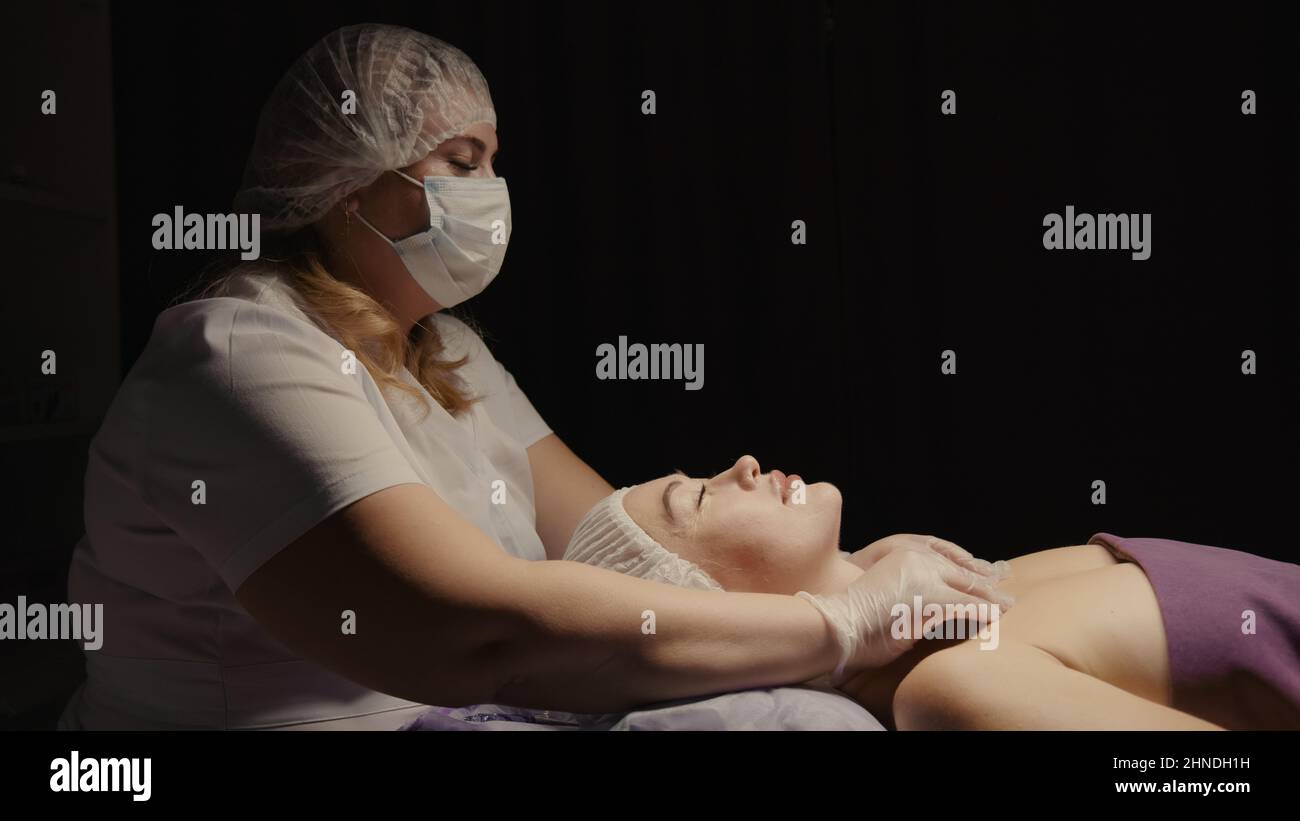 Junge hypnotische Gesichtsfrau erhält Anti-Aging-Gesichtsmassage im Spa-Salon Relax. Wellness Körper Hautpflege Gesicht Schönheitsbehandlung, Verjüngungskur bekommen Kopf Gesichtsmassage. Schwarzer Hintergrund Stockfoto