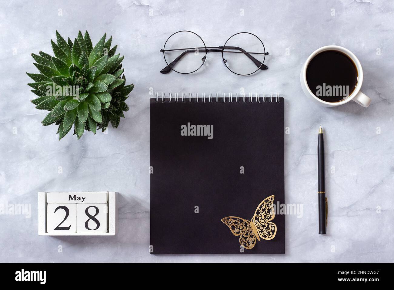 Schreibtisch im Büro oder zu Hause. Holzwürfel Kalender Mai 28. Schwarzer Notizblock, eine Tasse Kaffee, Sukkkulent, Gläser auf Marmorhintergrund Konzept stilvolle Workpla Stockfoto