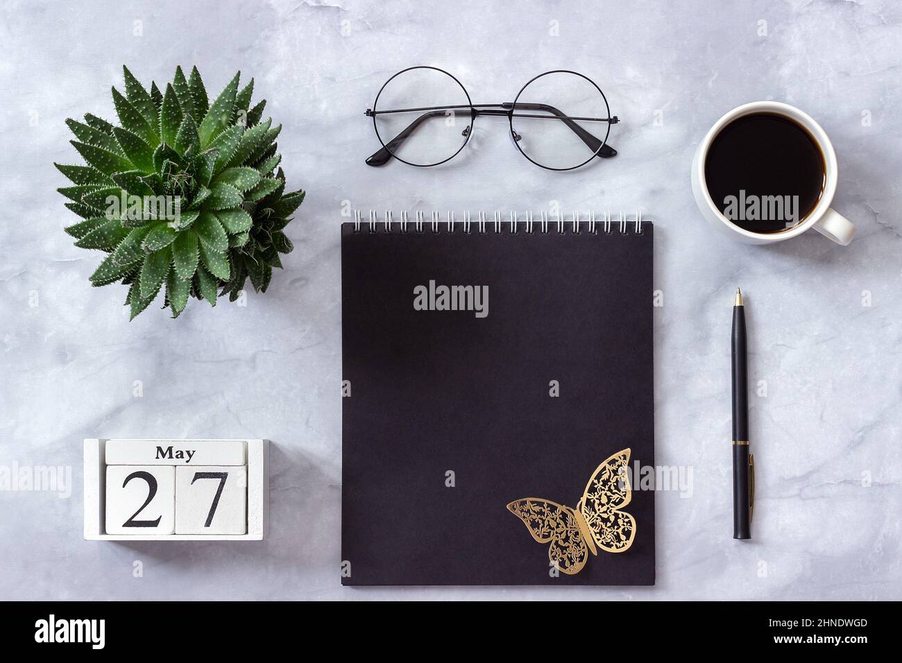Schreibtisch im Büro oder zu Hause. Holzwürfel Kalender Mai 27. Schwarzer Notizblock, eine Tasse Kaffee, Sukkkulent, Gläser auf Marmorhintergrund Konzept stilvolle Workpla Stockfoto