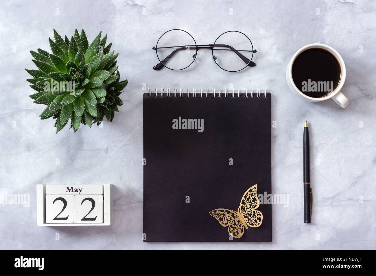 Schreibtisch im Büro oder zu Hause. Holzwürfel Kalender Mai 22. Schwarzer Notizblock, eine Tasse Kaffee, Sukkkulent, Gläser auf Marmorhintergrund Konzept stilvolle Workpla Stockfoto