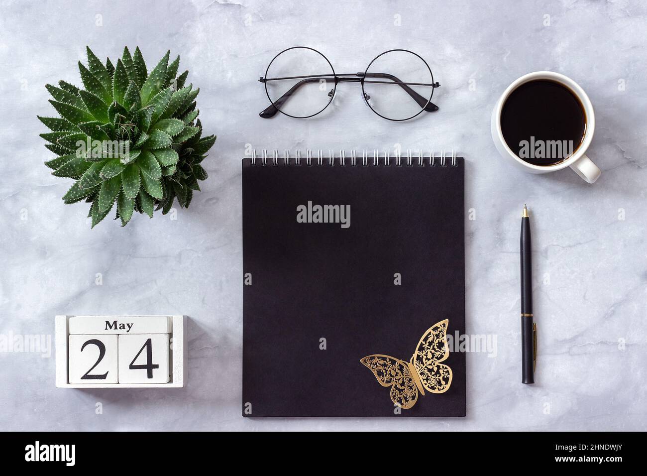 Schreibtisch im Büro oder zu Hause. Holzwürfel Kalender Mai 24. Schwarzer Notizblock, eine Tasse Kaffee, Sukkkulent, Gläser auf Marmorhintergrund Konzept stilvolle Workpla Stockfoto