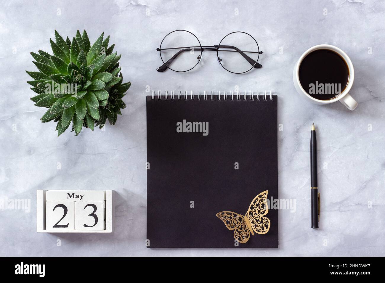 Schreibtisch im Büro oder zu Hause. Holzwürfel Kalender Mai 23. Schwarzer Notizblock, eine Tasse Kaffee, Sukkkulent, Gläser auf Marmorhintergrund Konzept stilvolle Workpla Stockfoto