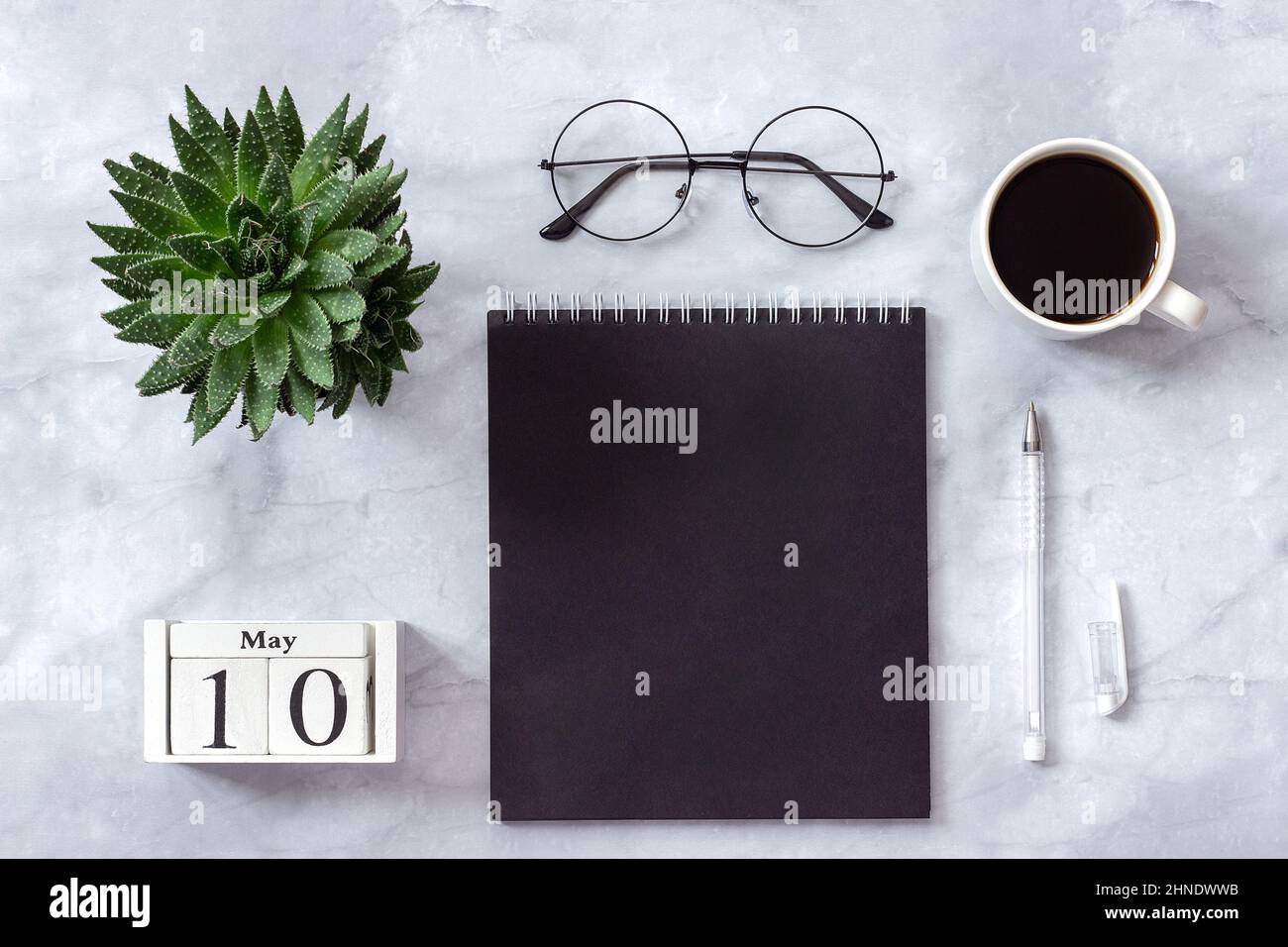 Schreibtisch im Büro oder zu Hause. Holzwürfel Kalender Mai 10. Schwarzer Notizblock, eine Tasse Kaffee, Sukkkulent, Gläser auf Marmorhintergrund Konzept stilvolle Workpla Stockfoto